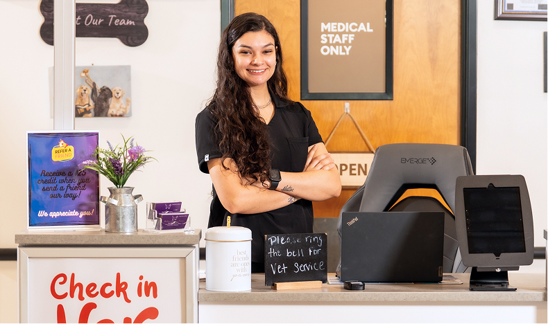 A veterinary staff member stands with arms crossed behind a reception desk. A sign in front of the desk reads, "Check in here."