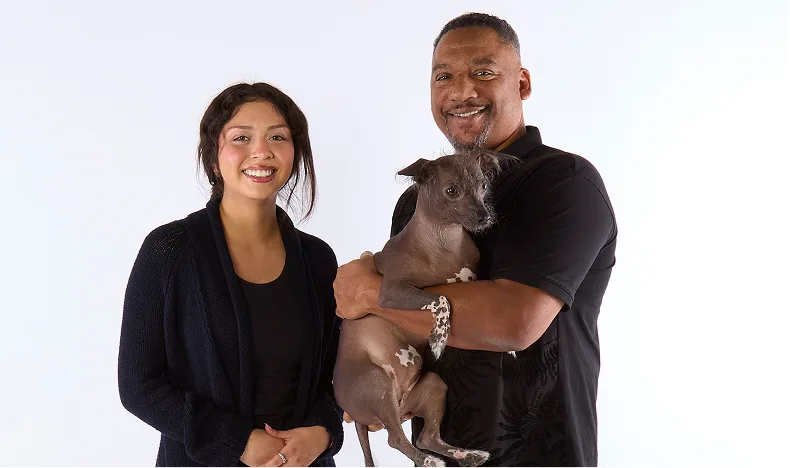 Two people pose together, one holding a dog in his arms.